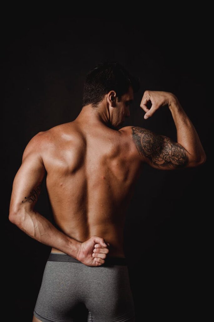 A shirtless muscular man flexes his biceps, captured from behind, displaying tattoos.